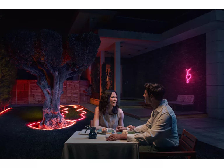 Two people sitting at a table in a dark outdoor setting with a Philips Hue Neon Outdoor Strip Light in the background.