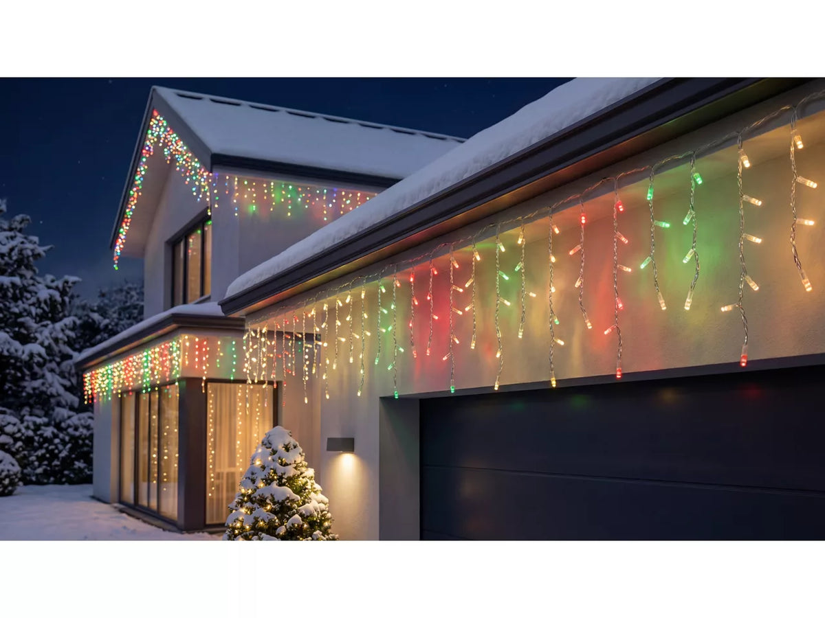 Multicolor Philips Hue Festavia Icicle Christmas string lights draped along the roofline and garage of a snow-covered house at night, with a small decorated tree in the foreground.