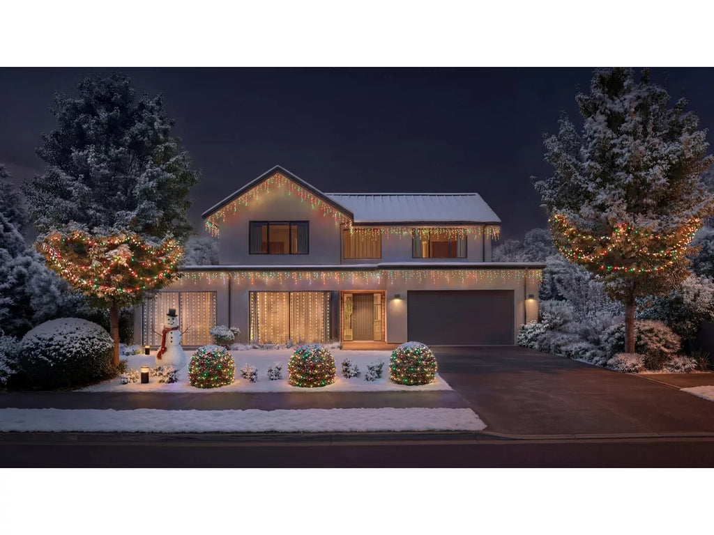 A modern single-story house at night covered in warm white holiday string lights, with snow on the roof and landscaping. Snow-covered trees and shrubs frame the front yard, and a snowman decoration stands near the left side of the porch with the Philips Hue Festavia Icicle Lights lighting up the house
