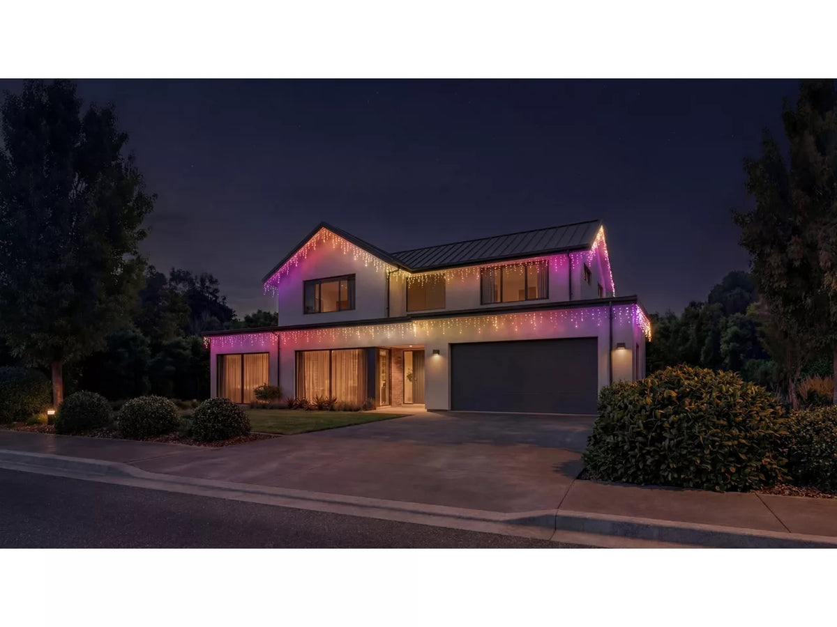 A modern two-story house at night with colorful string lights draped along the roofline and around the windows, casting a warm glow. The house has a dark gray garage door, concrete driveway, landscaped bushes, and trees on both sides under a dark night sky.