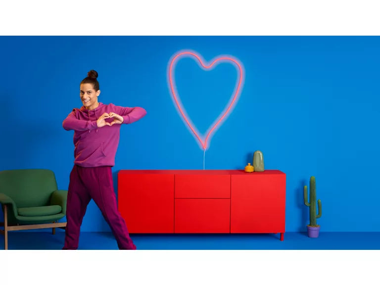 Person in purple outfit making heart shapes with hands, red cabinet, and blue wall with heart drawing.