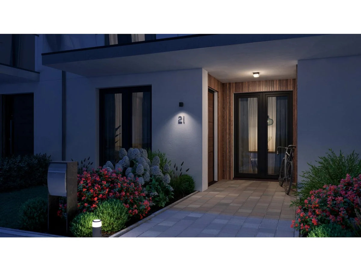 Modern front entrance at dusk with a Philips Hue Muscari wall spotlight lighting the wall beside the door and a garden path lined with flowers.