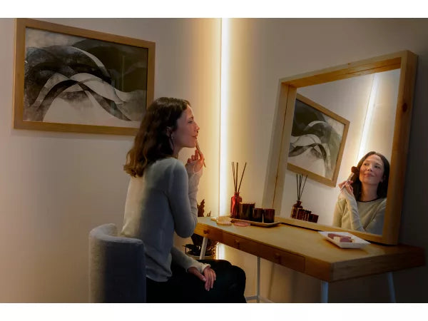 Woman sitting at a vanity table in a softly lit room with framed artwork on the wall and lighting from the Philips Hue OmniGlow Strip Light
