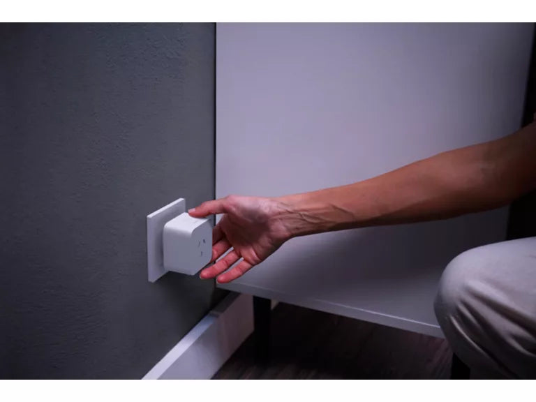 A person's hand reaching to plug or adjust a small white electronic device into a wall outlet, with a gray textured wall and white furniture visible.