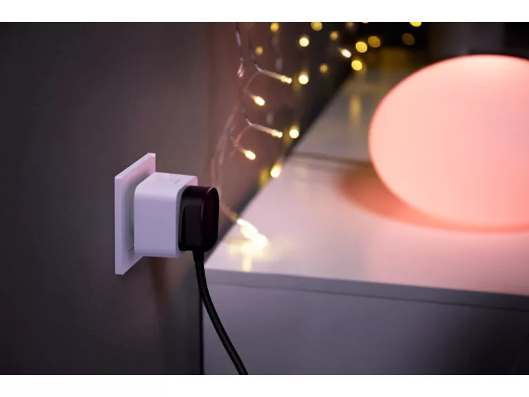 A white wall-mounted power adapter or plug with a black plug inserted, shown plugged into a wall beside a glowing pink orb lamp and warm string lights on a tabletop.
